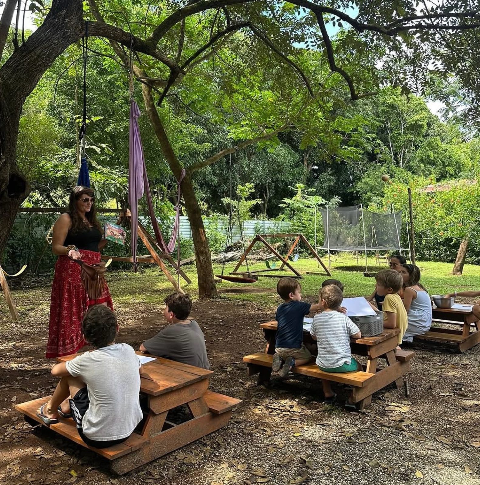 Children and parents gathered at outdoor picnic-table desks for a lesson under the trees at a local education setting