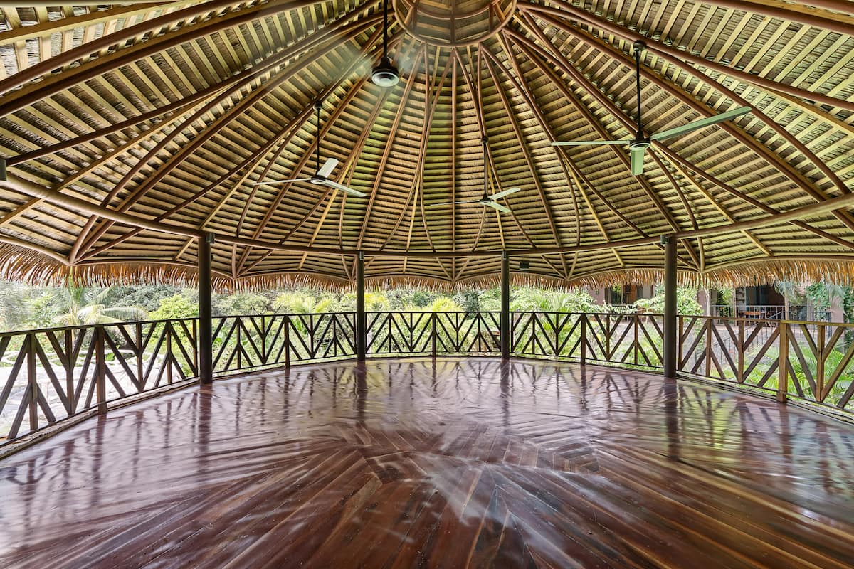 Inside the open-air yoga shala, polished hardwood floor under a soaring thatched roof, open to the jungle